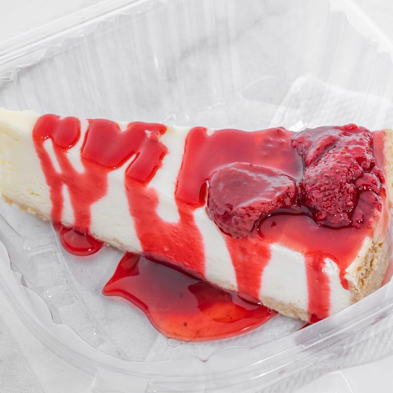 A slice of cheesecake with strawberry topping and whole strawberries in a plastic container.