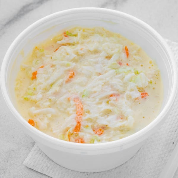 Creamy coleslaw with shredded cabbage and carrots in a white bowl on a white textured surface.